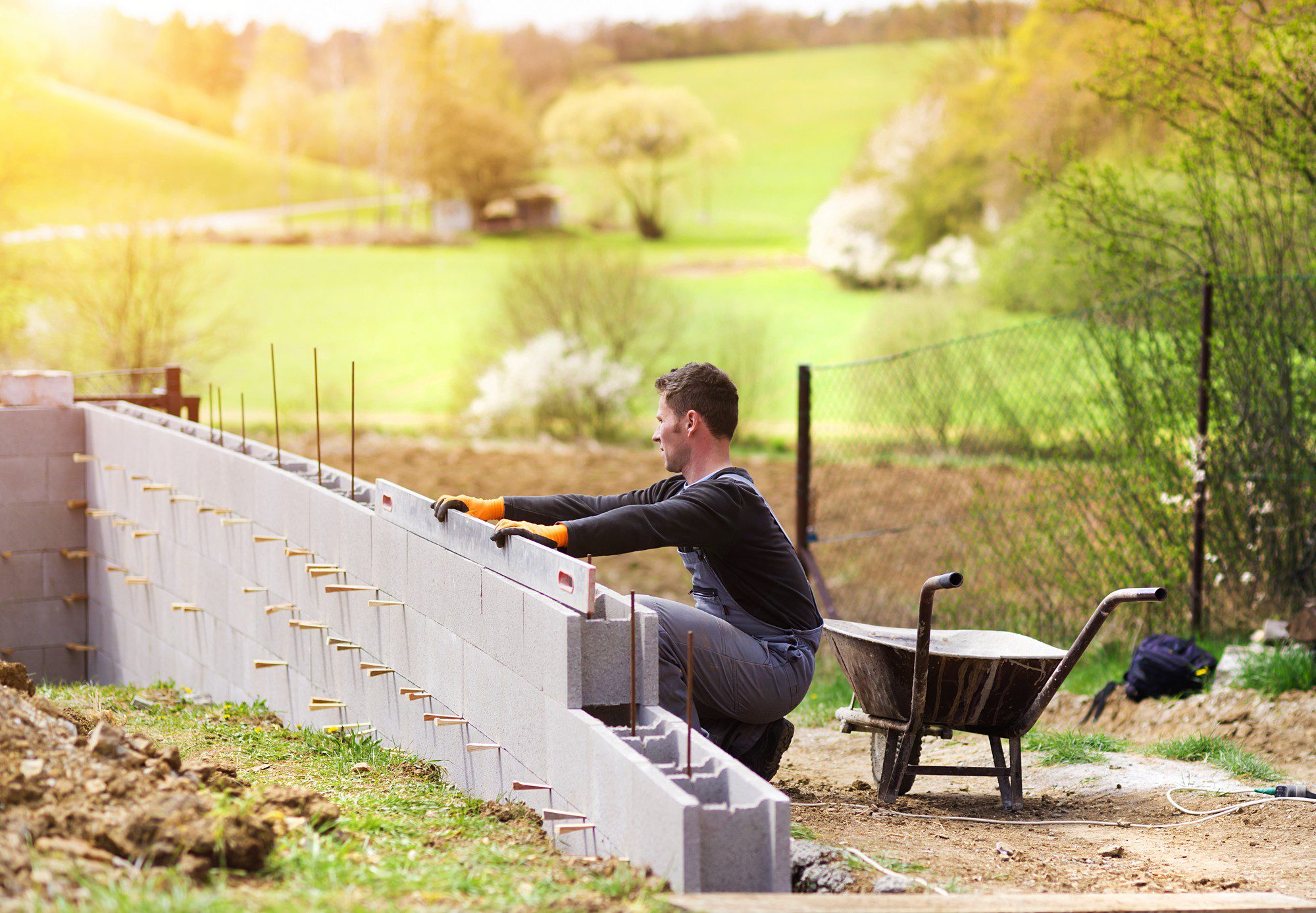 The image shows a construction setting where a person appears to be constructing a cinder block wall. The individual is wearing casual work attire with gloves, and is seated on the wall while examining or resting from the work. There's a wheelbarrow nearby, which is typically used to transport materials at a construction site. The environment around the construction area is grassy and rural with trees and fencing, suggesting the work is being done in a countryside setting. The weather seems to be fair, with sunlight illuminating the scene.