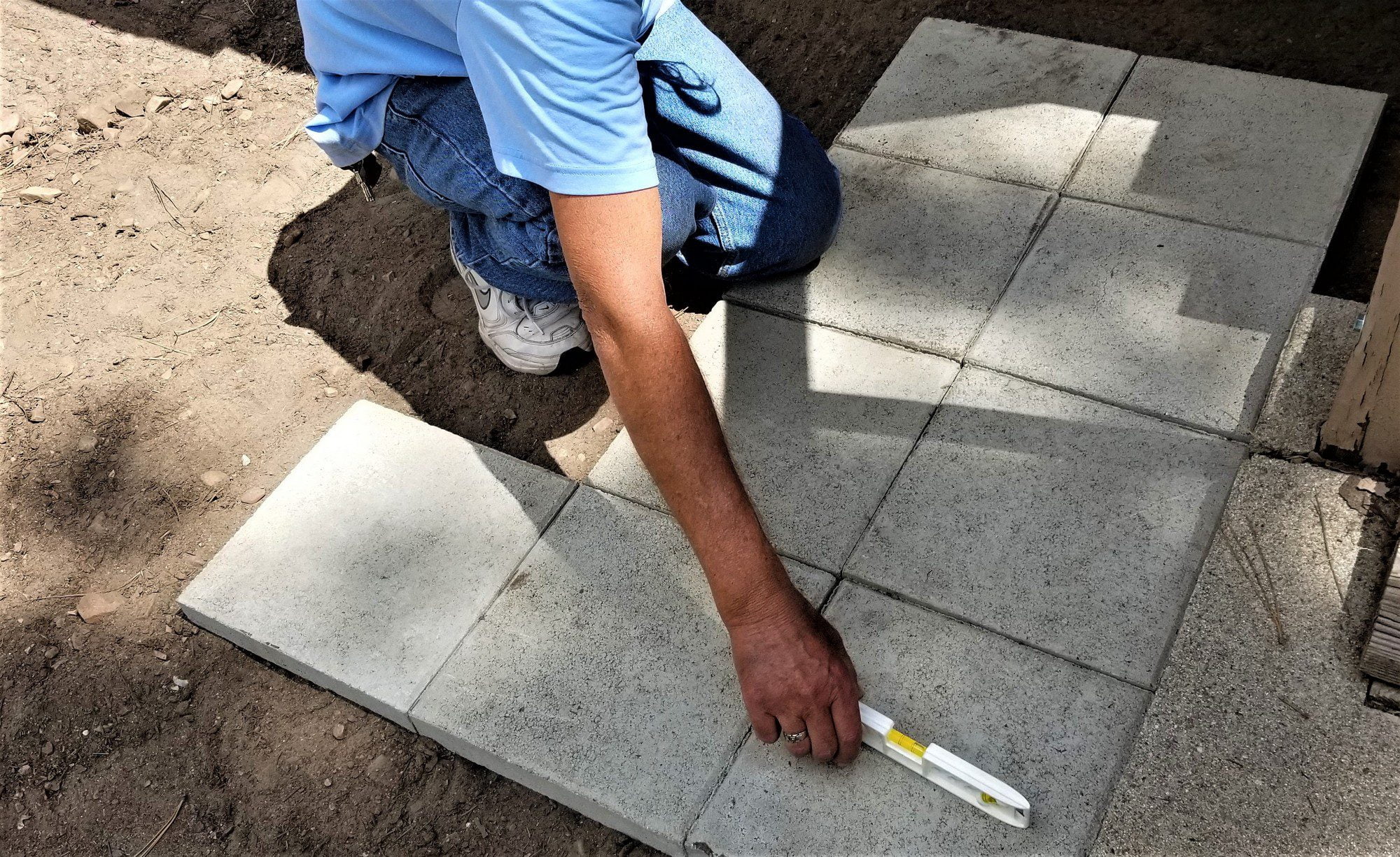 The image shows a person installing square concrete pavers or tiles outdoors. The person is bent over the work area and appears to be using a tool, possibly a rubber mallet or similar device, for setting the pavers in place. Some pavers are already laid down, forming a pattern on the ground. The photograph captures a moment of manual work, specifically the process of laying pavers to create a hard, flat surface, such as a patio, walkway, or driveway.
