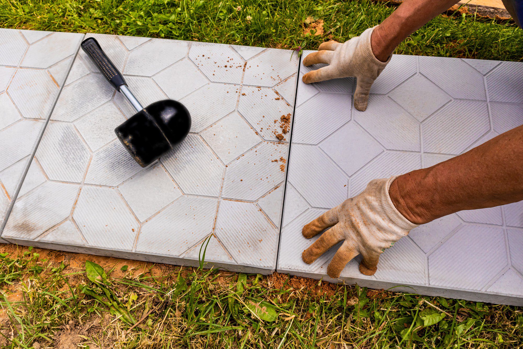 The image depicts a person laying interlocking tiles to create a pathway or patio area. The individual is wearing a pair of light-coloured work gloves, indicating some manual labour is being performed. In the upper left corner of the image, there is a rubber mallet, which is typically used in tile setting to gently tap tiles into place to ensure proper leveling and placement without cracking them. The tiles have a geometric, hexagonal design and appear to be light grey in colour. The grass around the installation area suggests this work is taking place outdoors, and the presence of soil and grass cuttings hint at recent groundwork preparation.