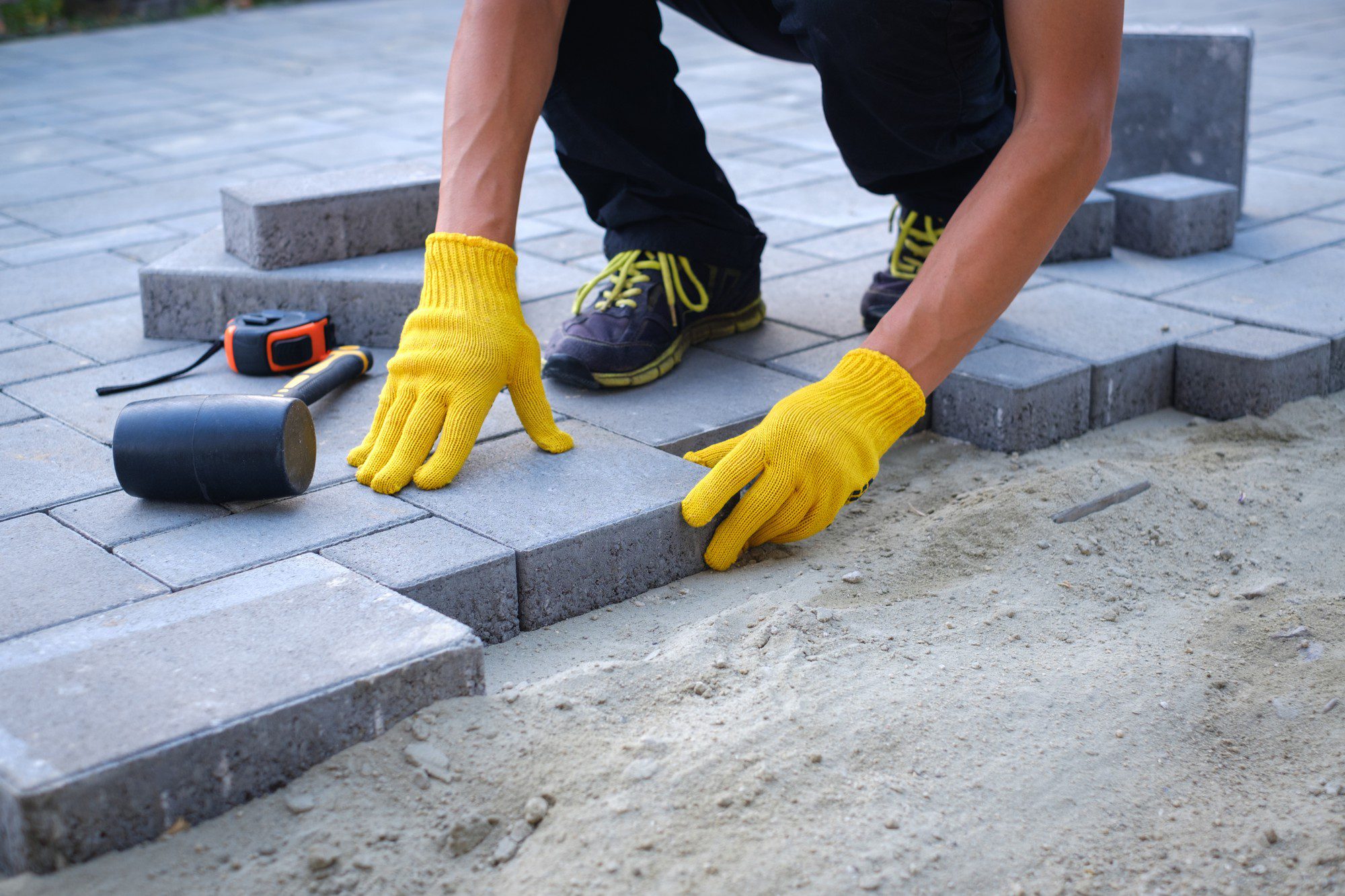 The image shows a person laying paving stones on a prepared sand base. The person is wearing yellow work gloves, and we can see them placing a paving stone into position on the ground. In the background, there are additional paving stones and a mallet, commonly used for tapping the stones into place, alongside a tape measure, which is likely used for accurate placement and spacing of the stones. The person is wearing casual sports shoes and seems to be engaged in outdoor construction or landscaping work. The focus is on the hands and the pavers, indicating the manual labour aspect of the task.